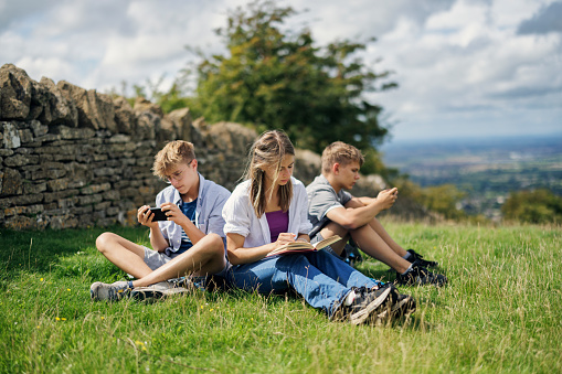 Three teenage kids are hiking in the Cotswolds, Worcestershire, United Kingdom. They enjoying a short break, sitting on a blanket and reading a book and using phones.
Overcast, summer day.
Shot with Canon R5