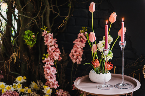 Table with pink candles, yellow flowers in vase, dry plant. Easter zone with rustic design, spring branches, black wall. Arch with luxury floral. Home decor for photo sessions. Birthday party detail.