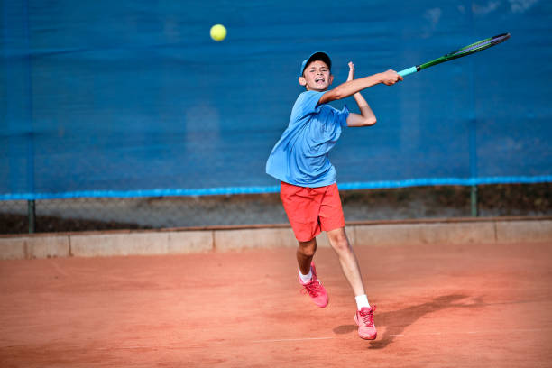 un jeune garçon caucasien joue au tennis sur un court orange en fin d’après-midi. - tennis child athlete sport photos et images de collection