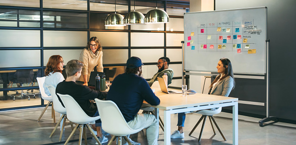 Business people in a boardroom engage in a creative discussion, exchanging ideas with a confident leader facilitating the meeting. The happy team represents successful collaboration and a modern tech company's productive work environment.
