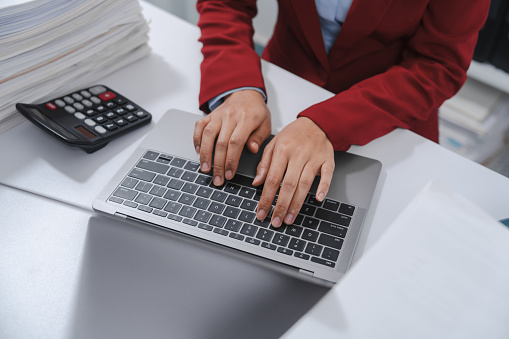 Close up hands of person in a red blazer using a calculator with one hand and typing on a laptop with the other, beside a large stack of paperwork.