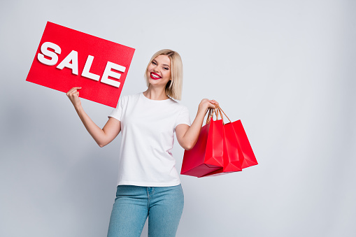 Photo of toothy beaming positive woman dressed white t-shirt hold shopping bags sale placard in arms isolated on white color background.