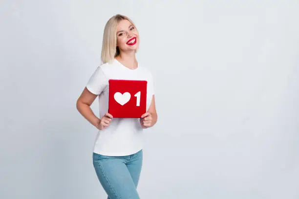 Photo of Portrait of toothy beaming lovely girl with blond hairdo wear t-shirt holding social media like in arms isolated on gray color background