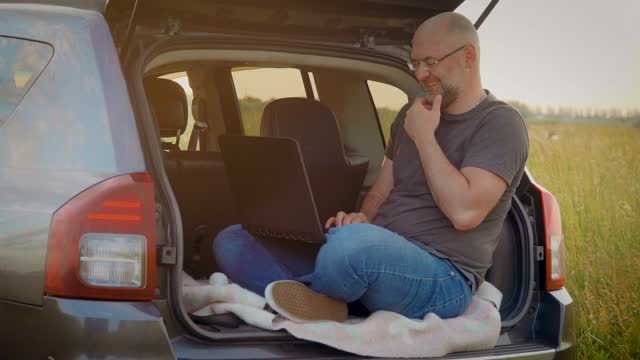 Happy smiling adult man enjoy workforce life hybrid digital work remotely anywhere sitting in car trunk while traveling