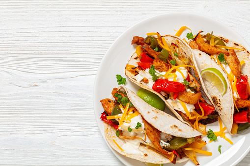 baked tex-mex chicken fajitas with mixed sweet pepper, onion, sour cream, cheddar cheese, lime, parsley and gluten free corn tortillas on plate on white wooden table, free space, flat lay, close-up
