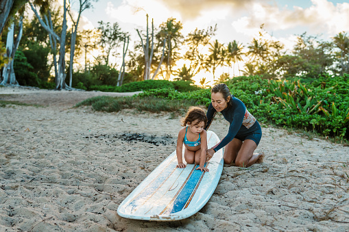 A determined four year old girl lays on a surfboard, practicing in the sand with the help of her active Hawaiian grandmother as she learns a new skill.