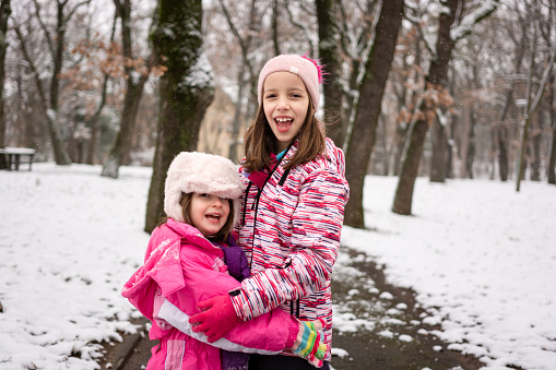 Portrait of two happy girls standing in each other's arms and looking at the camera during a snowy winter day in the park.