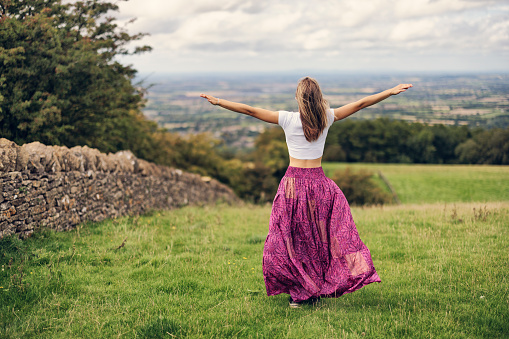 Teenage girl is hiking in the Cotswolds, Worcestershire, United Kingdom. She is standing on the beautiful meadow on top of the hill and looking at the view\nSunny and cloudy, summer day.\nShot with Canon R5