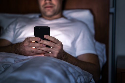 Phone in bed at night. Man with smartphone before sleeping. Secret texting, cheating or working late. Happy guy browsing online news. Holding cellphone in hand. Dark home bedroom. Screen time concept.