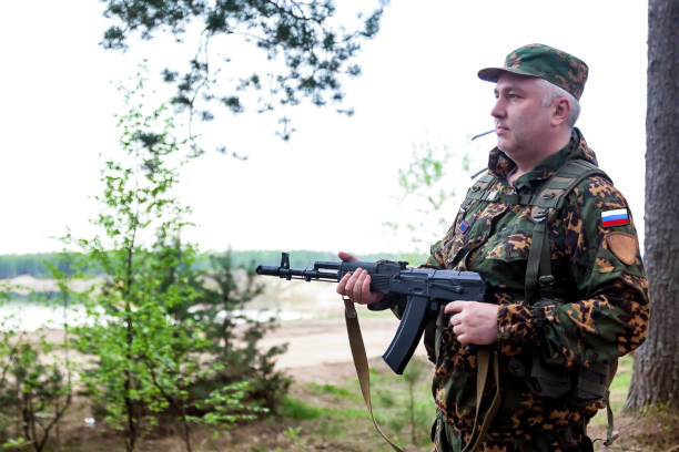 istock Russian soldier man dressed military camouflage uniform with weapon and cigarette in forest at clean sky background. Male border guard in country border with autogun on war. Copy text space 1922228921