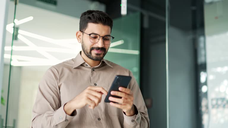 Smiling businessman is using smartphone standing in business office. Bearded employee reads or typing message,