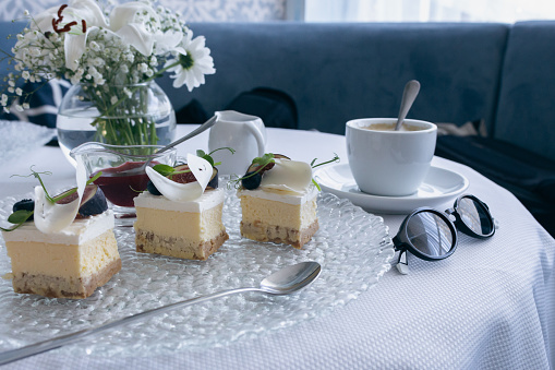 Cheesecake with cream and decor with cup of coffee, spoon and sunglasses. Sweet breakfast in restaurant. Pieces of fresh cheesecake. Bakery dessert. Tasty cake in cafe. Creamy dessert. Mascarpone pie.