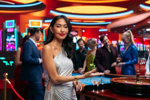 istock Advertising Template with an Asian Female Posing for Camera. Professional Beautiful Model Making a Hand Gesture and Pointing an Empty Template Space. Casino with People Gambling in the Background 1922377142