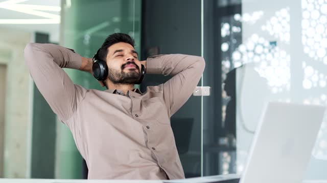 Bearded businessman in wireless headphones listening to relaxing music sitting with closed eyes at a workplace in a business office