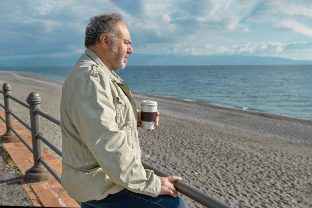 Pensive middle-aged bearded man drinking coffee on seaside  in sunny winter or spring day.