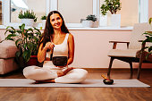 Happy beautiful young woman is sitting in lotus position and palyying singing bowl on yoga mat at yoga class. Soundhealing concept.