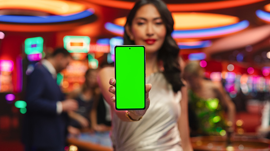 Advertising Template with an Asian Female Showing a Smartphone Device with a Green Screen Mock Up Display with Trackers. Professional Beautiful Model in a Casino with People Gambling in the Background