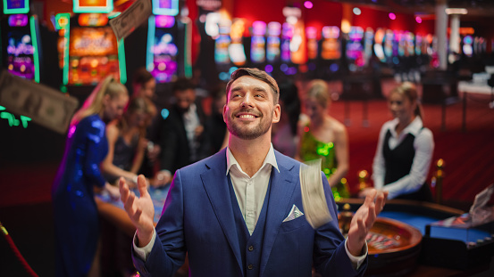 Happy Man in a Suit Standing in the Middle of a Modern Casino, Extending Arms, Welcoming a Money Rain.Young Male Smiling, Celebrating His Victory. Embodiment of a Successful Man