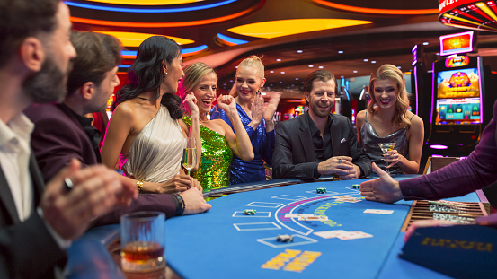 Diverse Group of Glamorous People Playing Game of Blackjack at Stylish Casino.Wealthy Patrons Having Good time Luxury Evening on Casino Floor, Placing Betts, Sociolizing. Gorgeous Lady Wins Jackpot