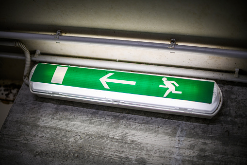 Vienna, Austria - Jan 29, 2020: Close up of illuminated emergency exit sign (colored green and white). Modern design, with running figure and arrow to the right. Neutral, white background.