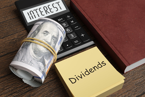 US dollar bankroll, calculator, notebook and a sticky note pad having the word Dividends written on the top on wooden table. Illustration of the concept of stock dividends