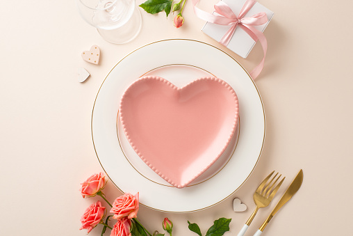 Intimate dinner affair with top view heart-shaped plates, cutlery, and wine glass. Beautiful roses, a gift box, and themed decor accentuate the pastel beige scene, inviting love and romance