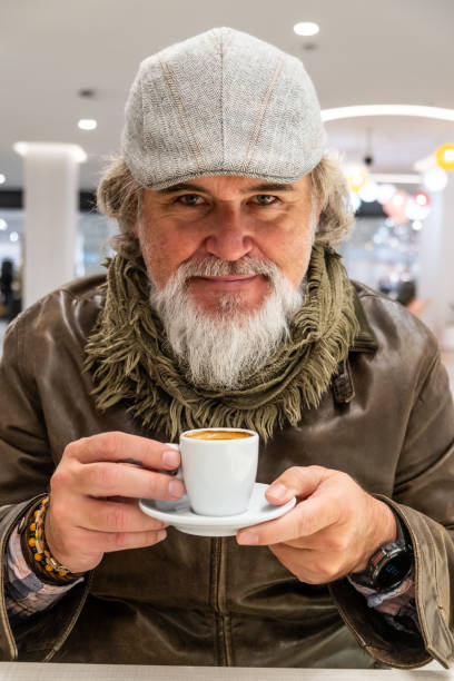 Middle-aged white bearded man in warm winter clothes holds a cup of hot coffee, looks at the camera and smiles.