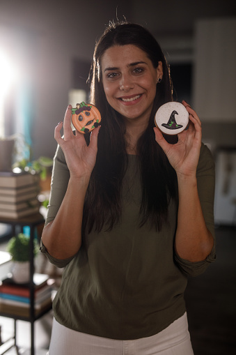 Portrait of charming young woman smiling at camera and showing off her Halloween themed cookies that she made.