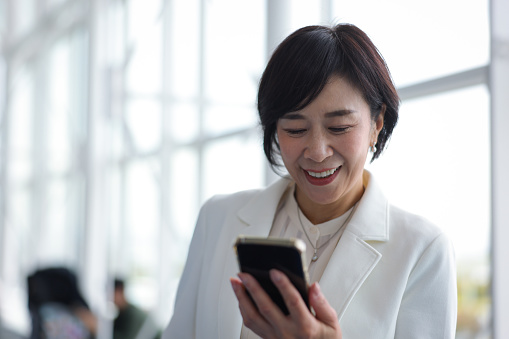 Smiling asian middle age woman with cell phone at international airport.
A female business owner or company officer goes on a business trip.
Airline ticket procedures are completed online on her mobile phone.
The woman purchased travel insurance and used a credit card at the airport before her trip.