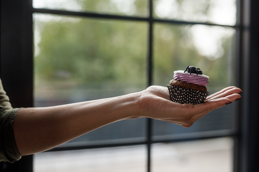 Close up shot of unrecognizable woman holding a cute Halloween themed cupcake in the palm of her hand against a window.