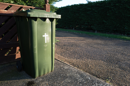 Green waste wheelie bin seen with a crucifix on the front. Located in an English cemetery. The full bin will be wheeled to the front of the cemetery for council collection.