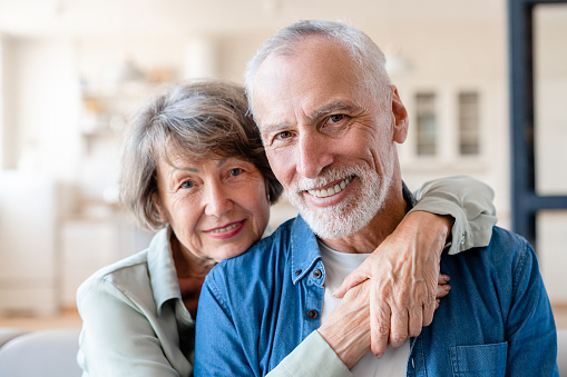 Senior old elderly spouses grandparents couple looking at camera while hugging embracing cuddling together at home. Physical touch - love language