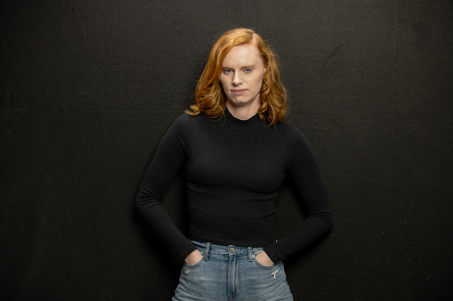A young woman in her early 20's is in a studio in Downtown Los Angeles, posing for the camera and enjoying her day. She wears a tight black sweater and faded jeans. He hair is  red and long and loose. She has freckles and a bright smile.