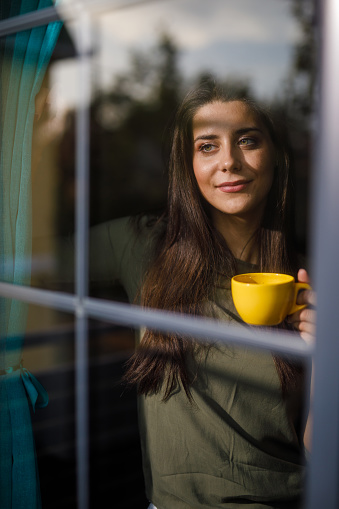 Portrait of happy young woman standing by the window in her home, enjoying a morning cup of coffee, looking outside, smiling and contemplating. Photographed through window.