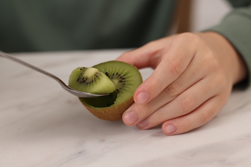 Boy eating tasty fresh kiwi with spoon at white marble table indoors, closeup