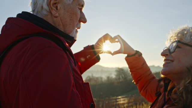 SLO MO Zoom Out Shot of Loving Senior Couple Making Heart Shape with Fingers and Kissing against Sunny Background