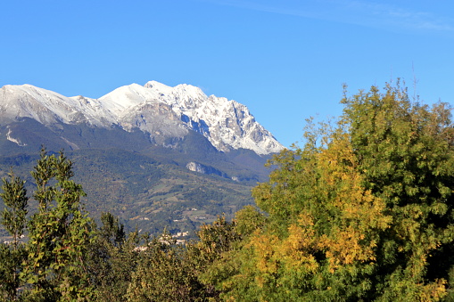 View of the Siella, Tremoggia and Camicia mountains, covered in snow on a sunny autumn day. At their foot, in the centre, above the tree line, the highest houses of the village of Farindola can be seen.