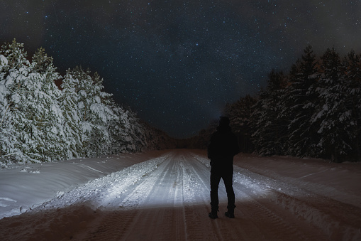Winter night scene, a man with a flashlight in a snowy forest on the road, starry sky with clouds. High quality photo