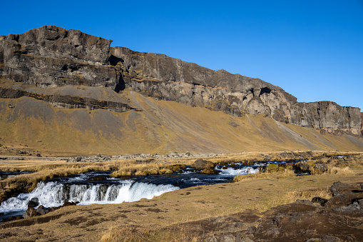 Mountain covered by moss and lichen, yellow in the autumn. Famous waterfall with its cascades. Bright blue sky. Fossalar, Iceland.