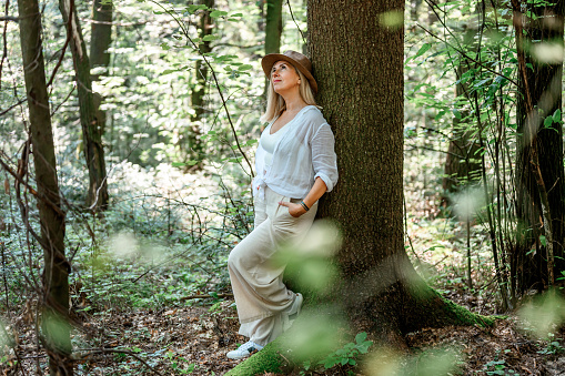 Adult blond woman in light summer clothes standing by the tree trunk in the woods and enjoying summertime.