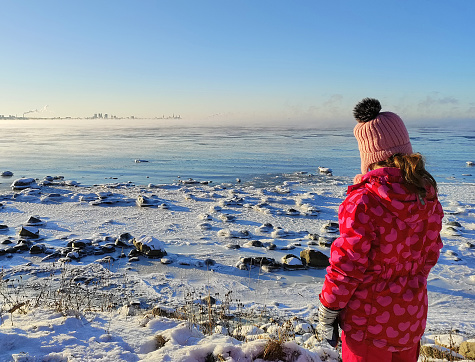 The child admires the sunny winter day and nature. Baltic sea covered with ice, Tallinn on the horizon.