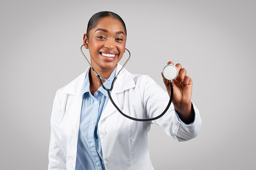 Smiling millennial african american woman doctor listening through stethoscope, diagnosis patient, medical exam, isolated on gray background, studio. Health care service, wellness