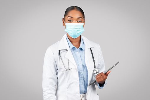 Smiling millennial black lady doctor therapist in white coat, protective mask hold tablet, isolated on gray background studio. Health care app remotely, consultation, exam