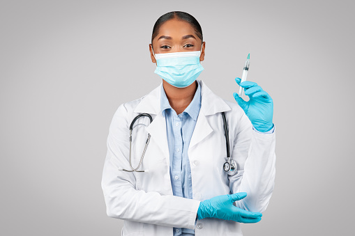Positive confident young black lady doctor in white coat, protective mask show syringe, recommends vaccination, isolated on gray background studio. Medicine and injection, health care in clinic