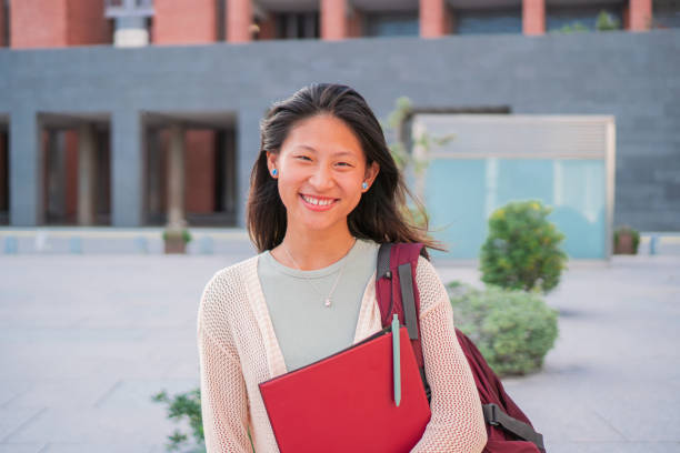 istock Young chinese student smiling and looking at camera holding a notebook and getting a backpack in her shoulder is standing individual and confidence with lovely smile outside the campus 1922985263