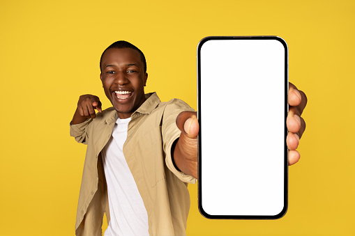 Glad excited young black man in casual pointing finger at mobile phone with empty screen, isolated on yellow studio background. Ad and offer, win, app for sale, website recommendation
