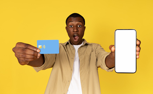 Shocked glad millennial african american guy show phone with empty screen and credit card, isolated yellow studio background. Ad and offer, app recommendation, banking and sale, online shopping