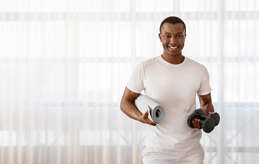 Cheerful handsome millennial black sportsman posing with yoga mat, barbells in his hands, smiling at camera, standing next to window, blank space for advertisement. Fitness at home