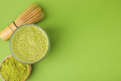 Glass of tasty matcha smoothie, powder and bamboo whisk on green background, flat lay. Space for text