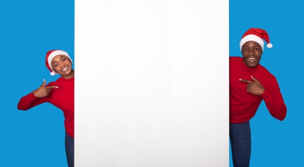 istock Black Couple In Santa Hats Pointing At Poster, Blue Background 1923100856
